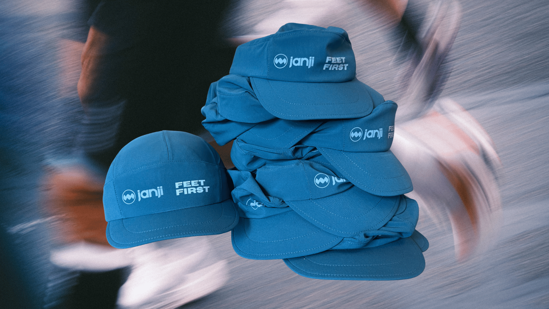 A stack of blue baseball caps with white embroidered logos and the words 'Janji FEET FIRST' is shown against a blurred background of people in motion.